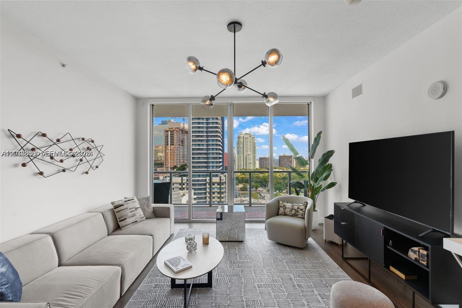 A modern living room features a sofa, armchair, television, and a balcony with city views — 79 SW 12th St, Miami FL 33130, MIAMI, FL