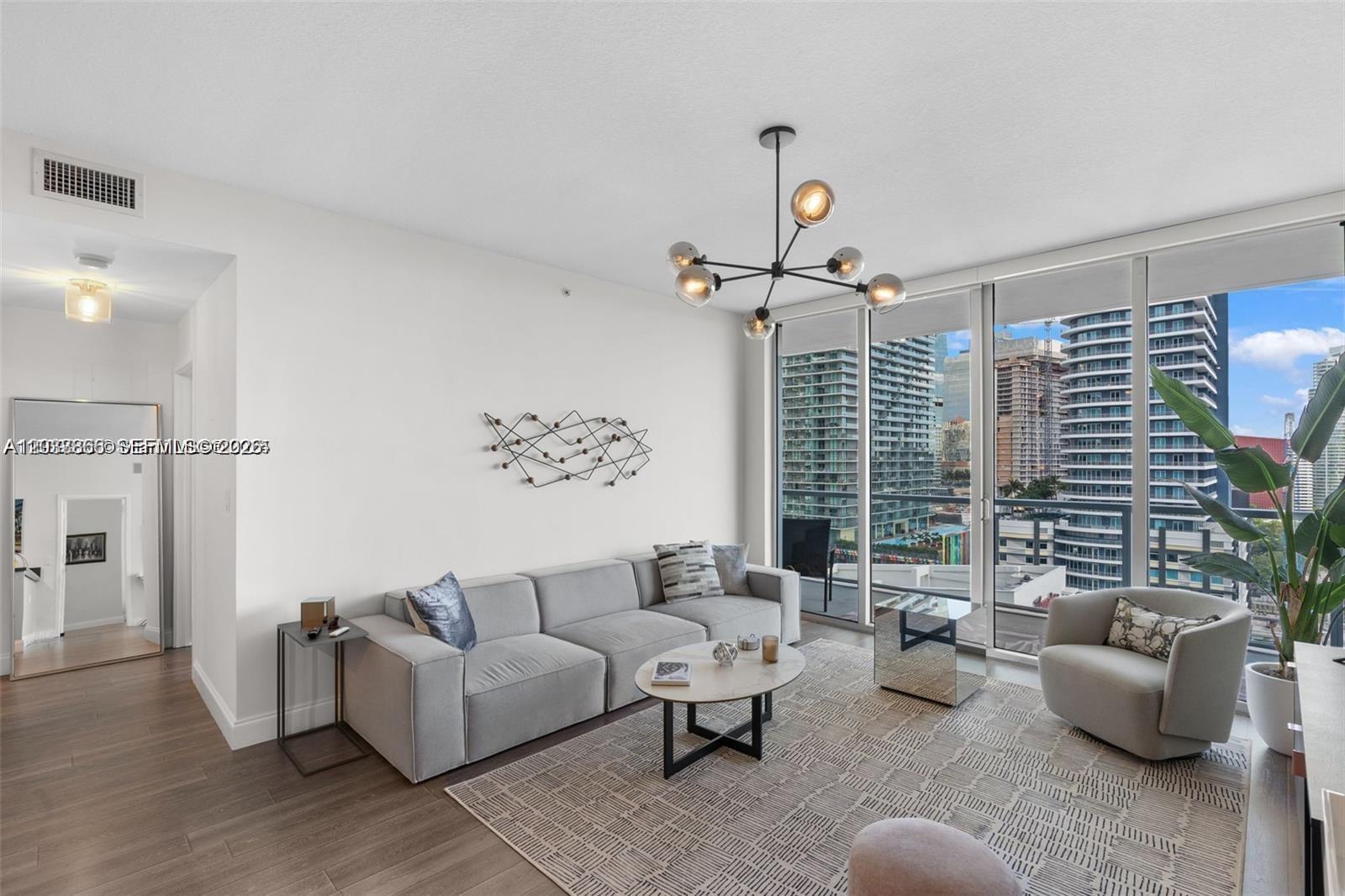 A modern living room features a gray sofa, patterned rug, and floor-to-ceiling windows overlooking buildings — 79 SW 12th St, Miami FL 33130, MIAMI, FL