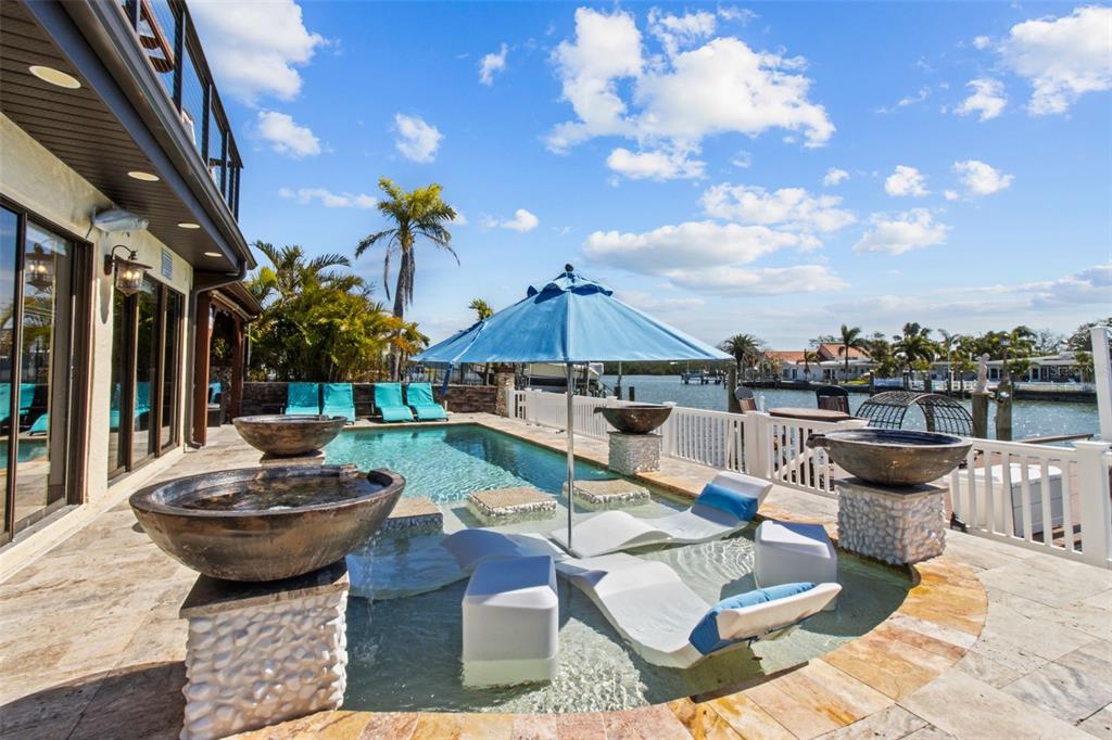 A waterfront patio features a swimming pool with lounge chairs and decorative stone water bowls — 357 LA HACIENDA DRIVE, INDIAN ROCKS BEACH, FL