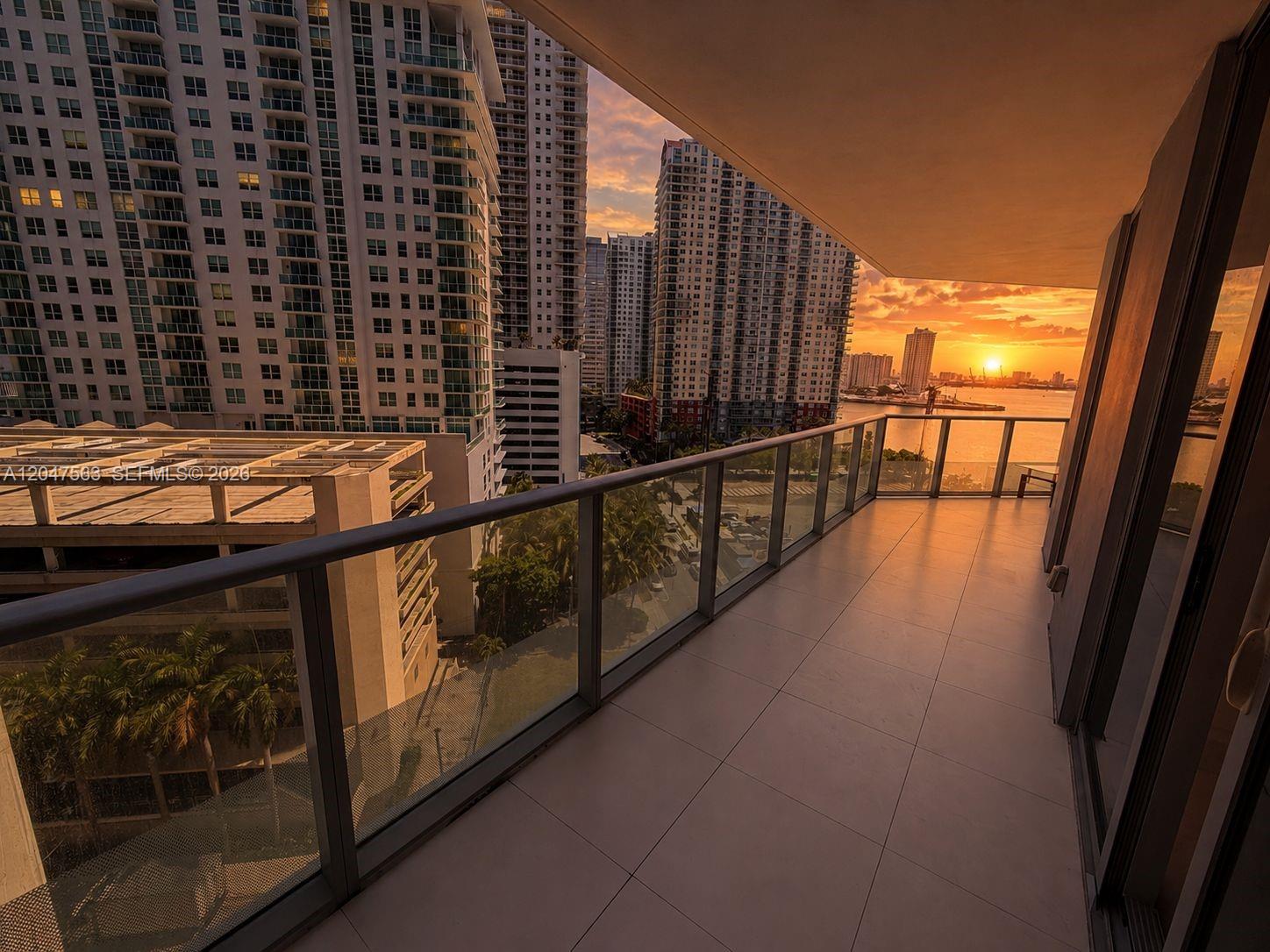A balcony with glass railings overlooks a city skyline and water at sunset — 1300 Brickell Bay Dr # 1110, Miami FL 33131, MIAMI, FL