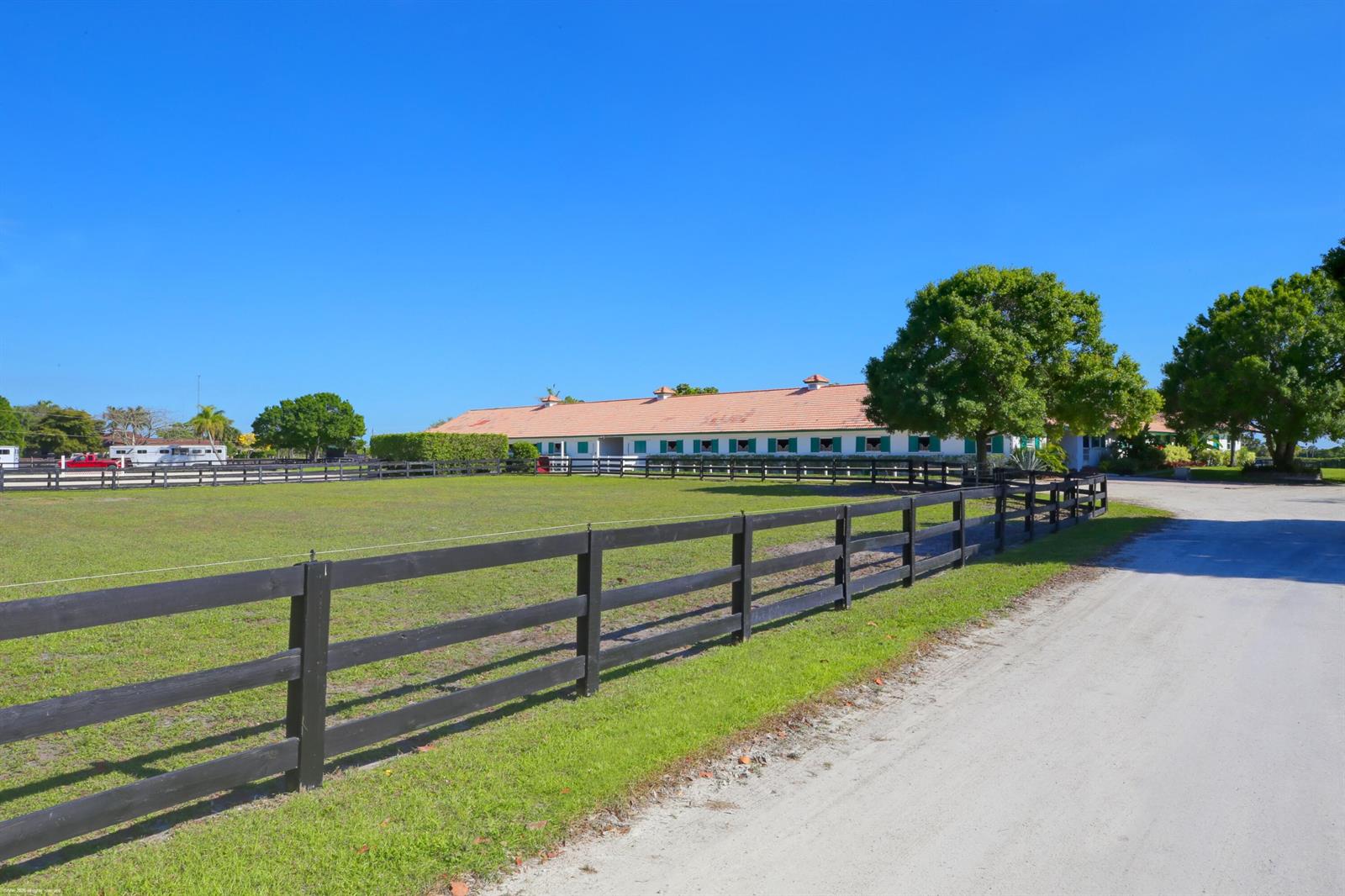 A long, single-story stable building sits behind a black wooden fence and grassy pasture — 13122 52nd Place S, Wellington FL 33449, WELLINGTON, FL