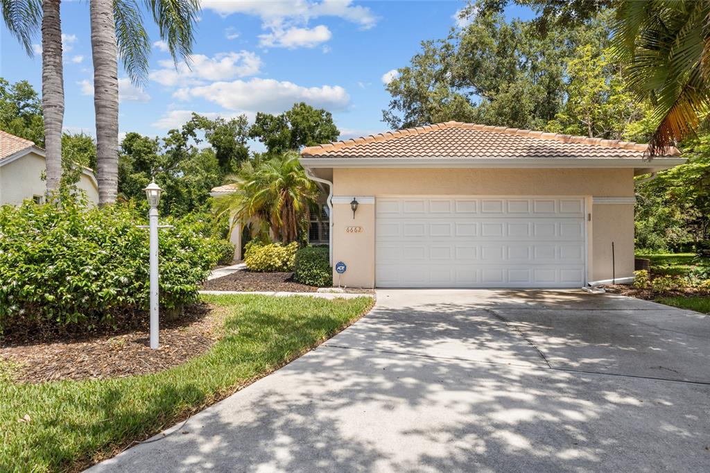 A beige house with a white garage door and a tiled roof sits behind a driveway — 6662 OAKBROOKE CIRCLE, BRADENTON, FL