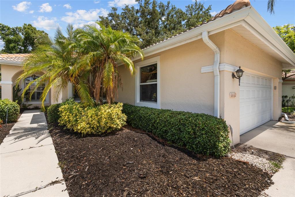 A beige house exterior features a palm tree, a front walkway, and a garage door — 6662 OAKBROOKE CIRCLE, BRADENTON, FL