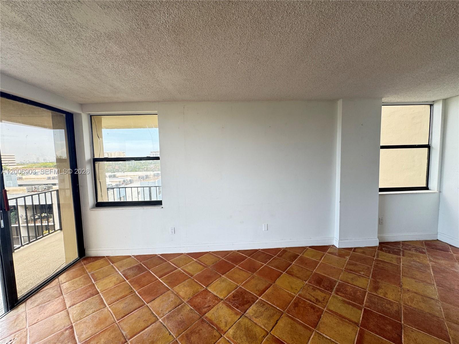 An empty room features terracotta tile flooring, white walls, and windows overlooking a city view — 1470 NE 123rd St # A701, North Miami FL 33161, NORTH MIAMI, FL