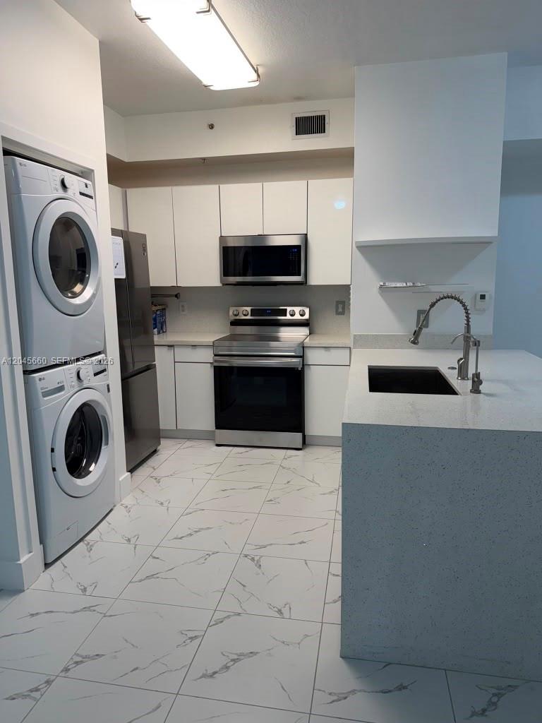 A modern kitchen features white cabinets, stainless steel appliances, a stacked washer-dryer, and marble-patterned flooring — 7220 NW 114th Ave # 105, Doral FL 33178, DORAL, FL