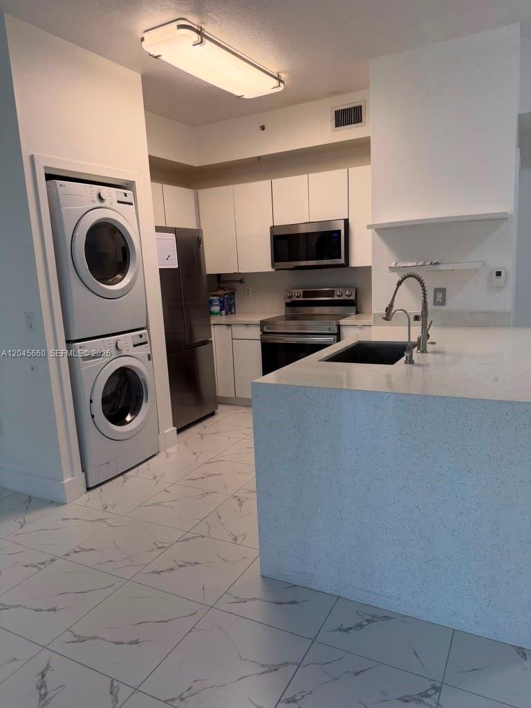 A modern kitchen features white cabinetry, stainless steel appliances, a stacked washer-dryer, and marble-patterned flooring — 7220 NW 114th Ave # 105, Doral FL 33178, DORAL, FL
