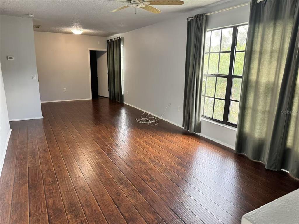 An empty living room features dark wood-look flooring, light gray walls, and large windows with curtains — 3366 MERMOOR DRIVE UNIT 206, PALM HARBOR, FL