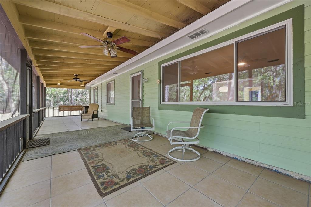 A long, covered porch features light green siding, tiled flooring, and several outdoor chairs — 8856 NW 230TH STREET, MICANOPY, FL