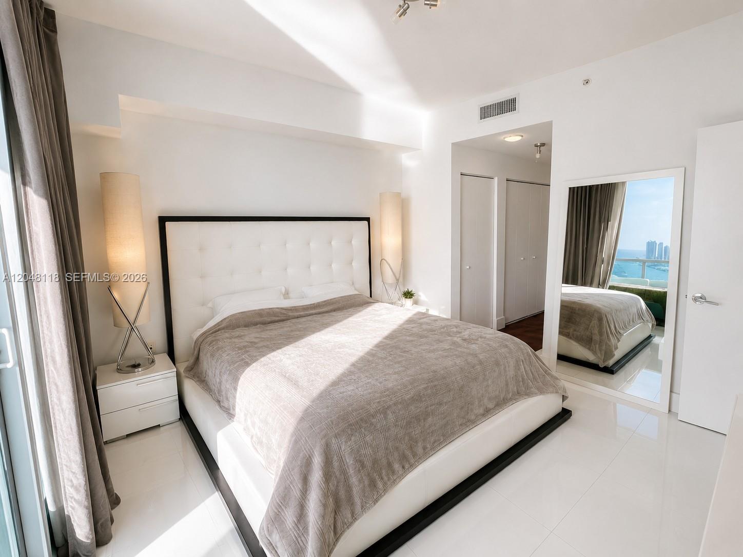 A modern bedroom features a tufted white headboard, a large mirror, and ocean views — 900 Biscayne Blvd # 5505, Miami FL 33132, MIAMI, FL