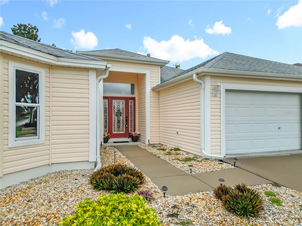 A tan house exterior features a red front door, a garage, and a gravel yard — 16946 SE 93RD CUTHBERT CIRCLE, THE VILLAGES, FL