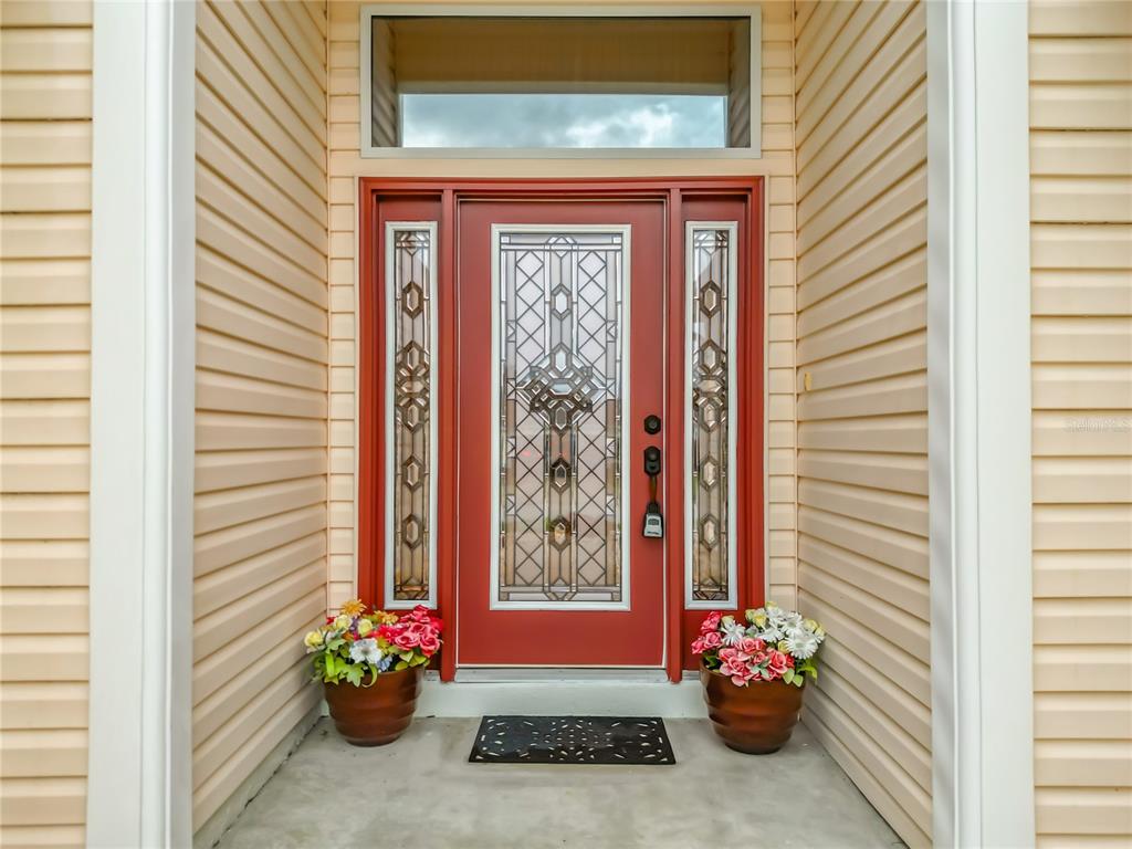 A red front door with decorative glass panels is flanked by two potted flower arrangements — 16946 SE 93RD CUTHBERT CIRCLE, THE VILLAGES, FL