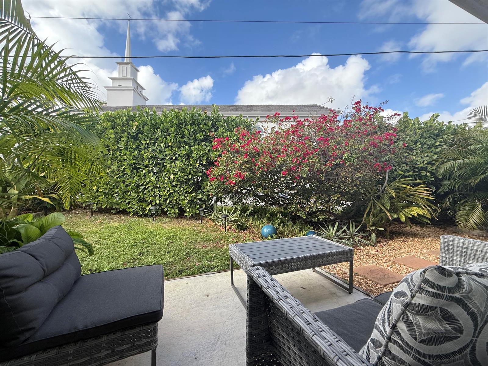 A patio with wicker furniture overlooks a grassy yard, flowering bushes, and a church steeple — 203 Seacrest Lane # A, Delray Beach FL 33444, DELRAY BEACH, FL