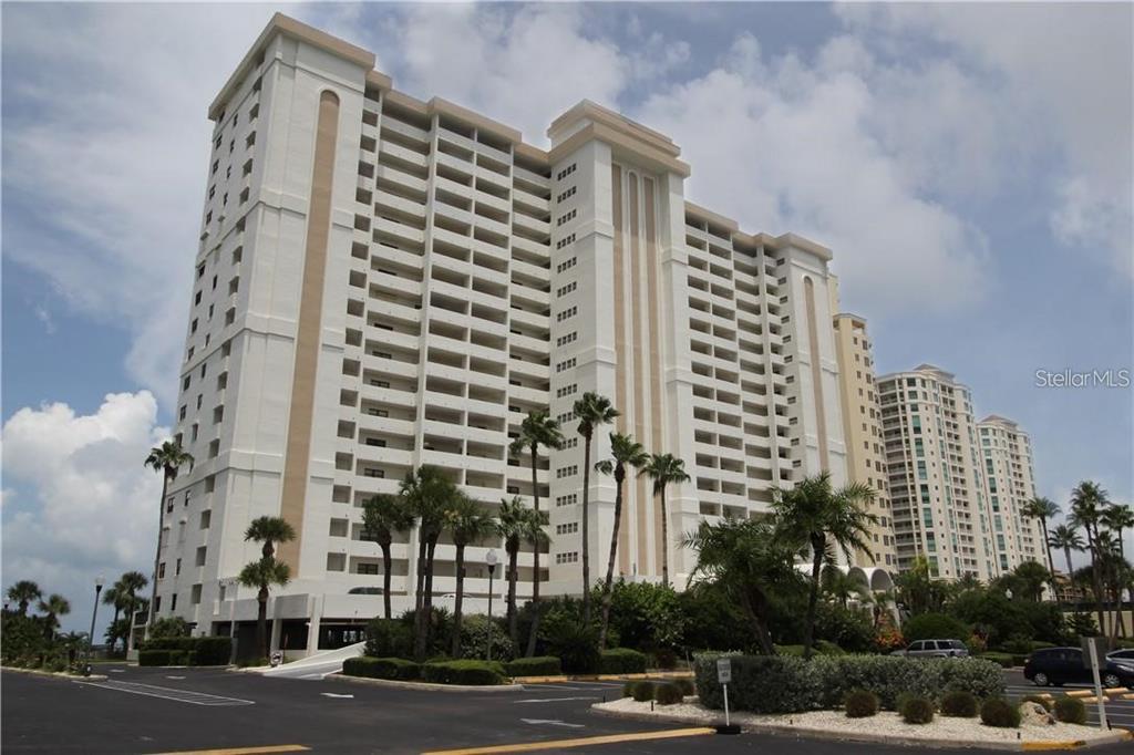 A tall, multi-story white residential condominium building stands behind a parking lot and palm trees — 1230 GULF BOULEVARD UNIT 1805, CLEARWATER, FL
