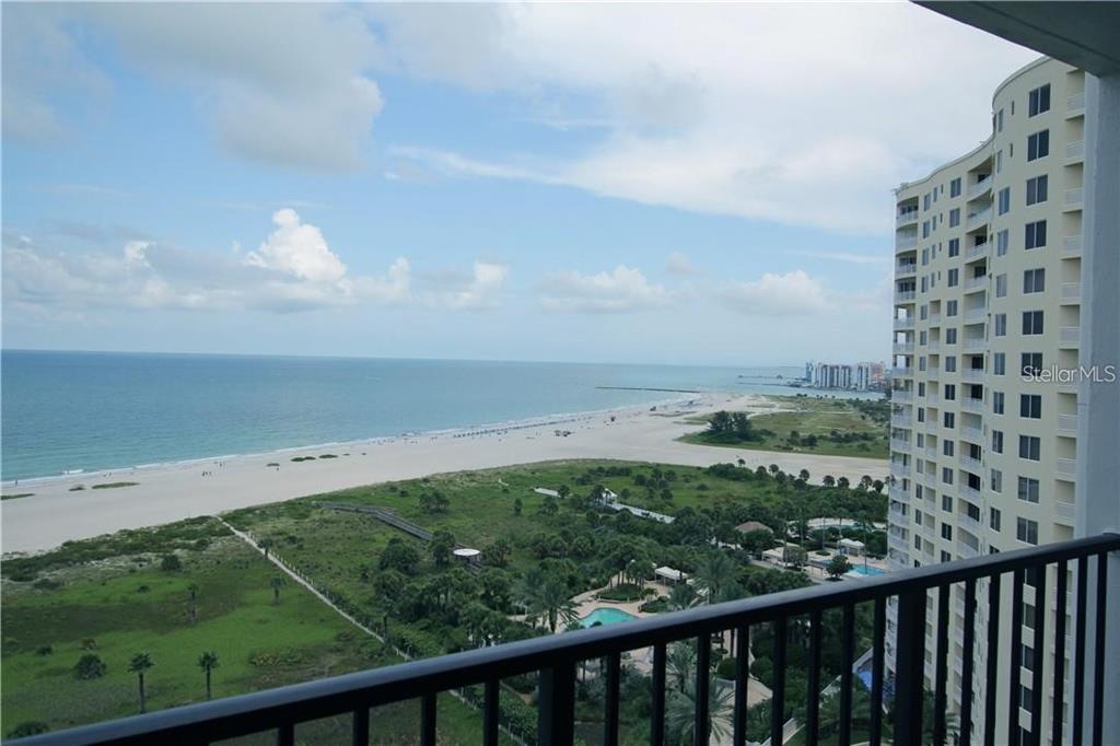 A high-angle view from a balcony overlooking a sandy beach, ocean, and a tall building — 1230 GULF BOULEVARD UNIT 1805, CLEARWATER, FL