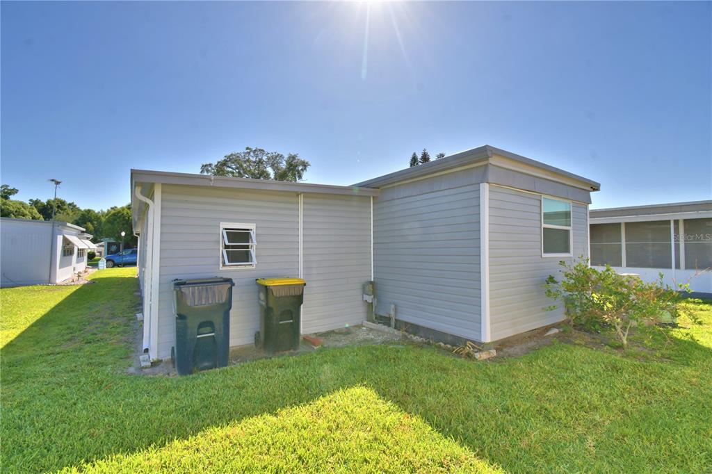 The exterior of a light gray mobile home features two trash bins and a small window — 117 HOLIDAY LANE, AUBURNDALE, FL