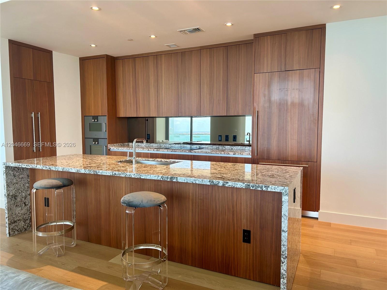 A modern kitchen features wood cabinetry, a granite waterfall island, and a mirrored backsplash — 175 SE 25th Road # 2804, Miami FL 33129, MIAMI, FL