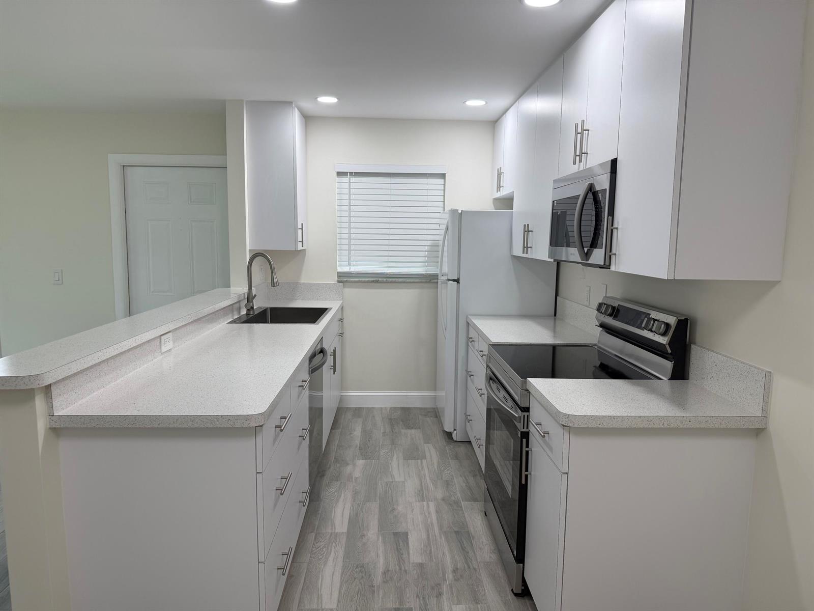 A galley kitchen features white cabinets, speckled countertops, stainless steel appliances, and grey wood flooring — 255 South Boulevard # D, Boynton Beach FL 33435, BOYNTON BEACH, FL