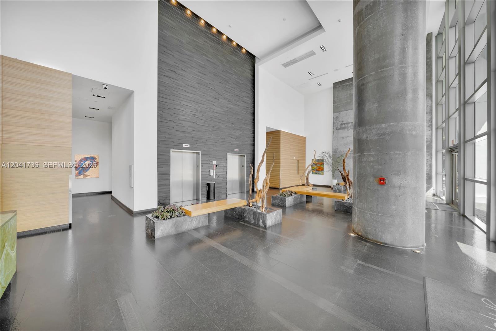 A modern building lobby features a large concrete column, two elevators, and wooden benches — 888 Biscayne Blvd # 2101, Miami FL 33132, MIAMI, FL