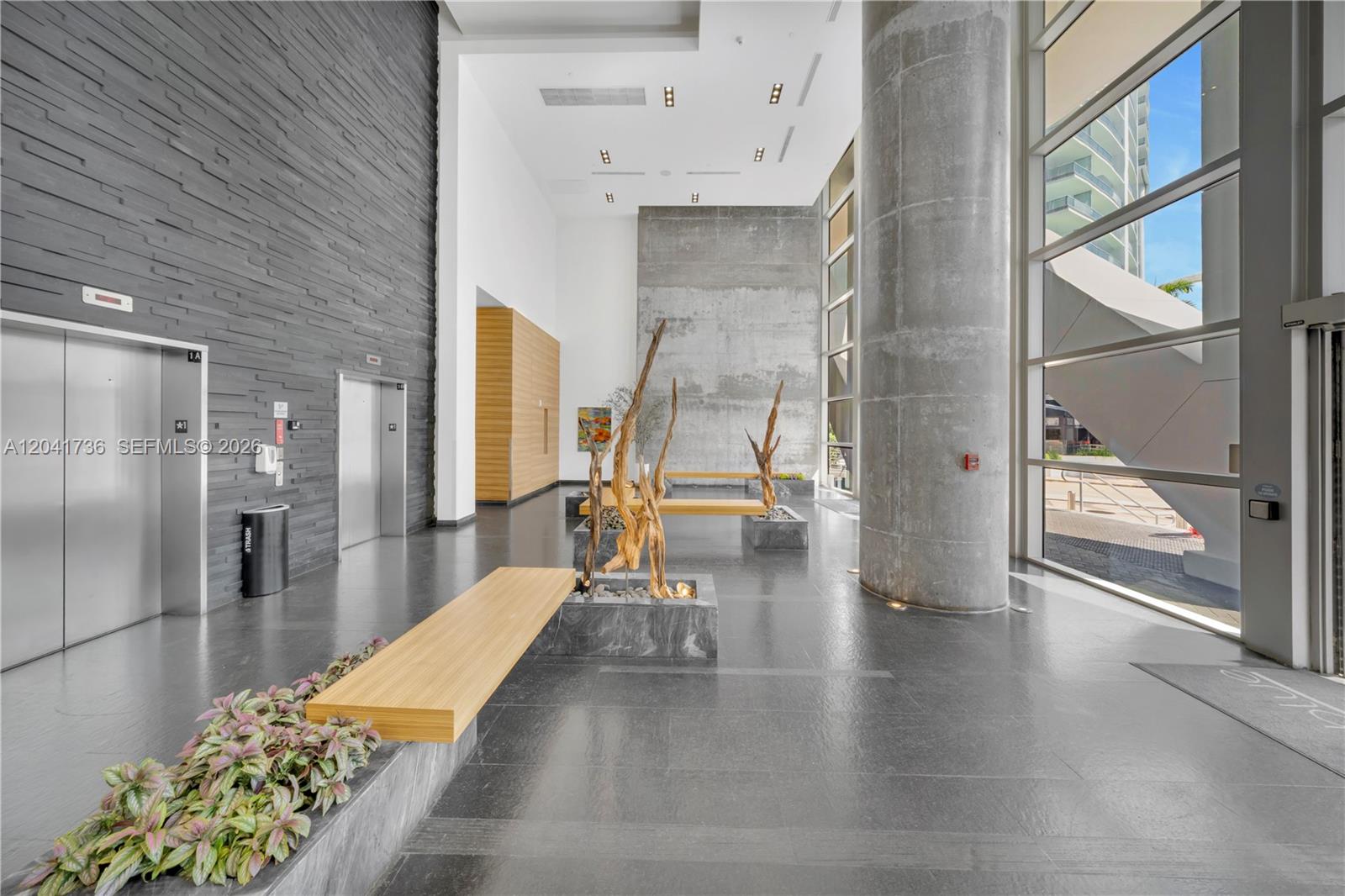 A modern building lobby features gray stone walls, a large concrete column, and wooden benches — 888 Biscayne Blvd # 2101, Miami FL 33132, MIAMI, FL
