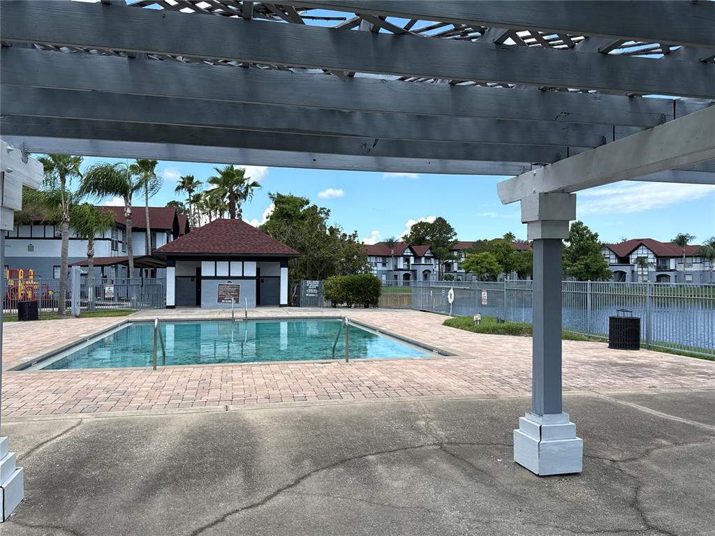 A swimming pool sits on a brick patio beneath a wooden pergola near apartment buildings — 3725 CASTLE PINES LANE UNIT 4322, ORLANDO, FL