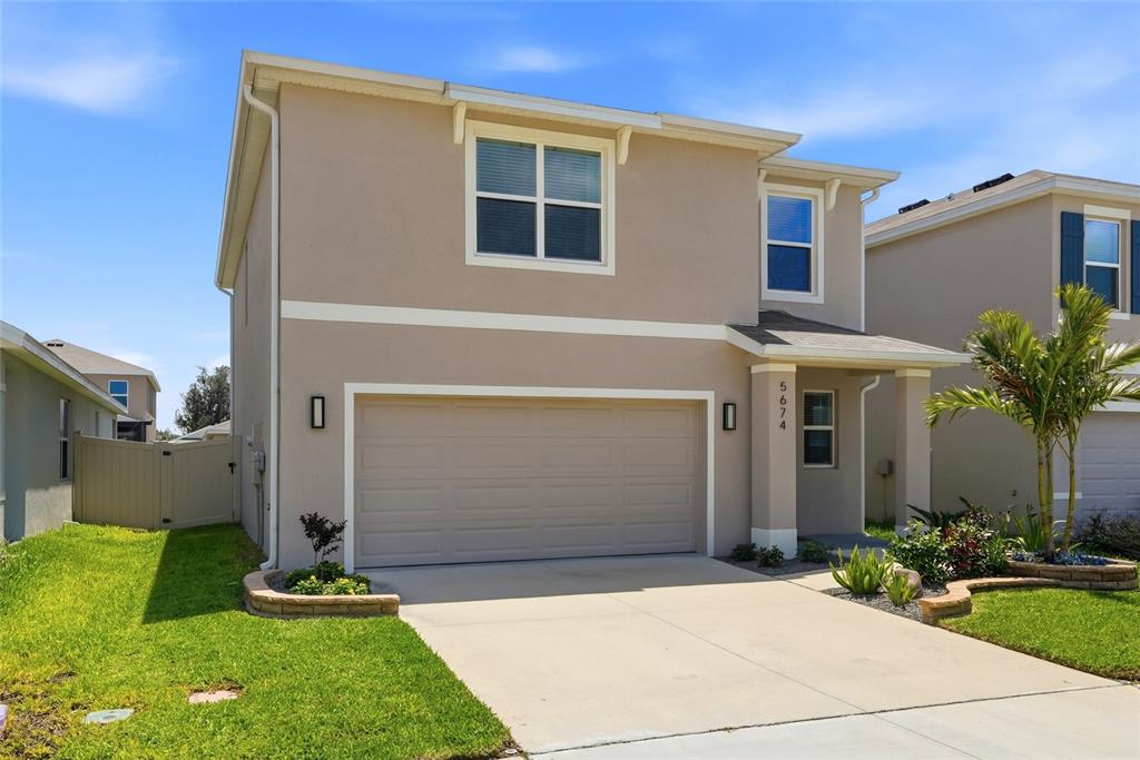 A two-story suburban house features a tan stucco exterior, a two-car garage, and landscaping — 5674 CHORLEYWOOD LANE, WESLEY CHAPEL, FL