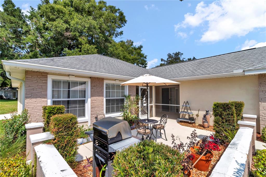 A patio with a grill, outdoor furniture, and an umbrella sits beside a brick-accented house — 7229 SW 115TH PLACE, OCALA, FL