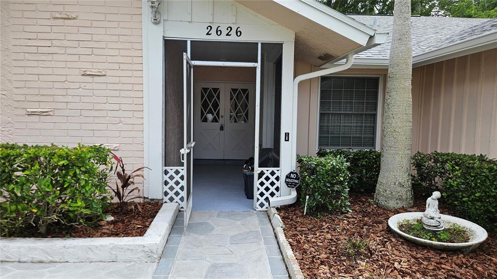 The exterior entrance features a screened-in porch, a stone walkway, and a small garden statue — 2626 WENDOVER TERRACE, PALM HARBOR, FL