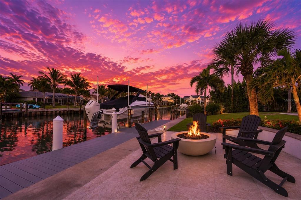 A waterfront patio features a fire pit, Adirondack chairs, and a boat docked at sunset — 465 E ROYAL FLAMINGO DRIVE, SARASOTA, FL