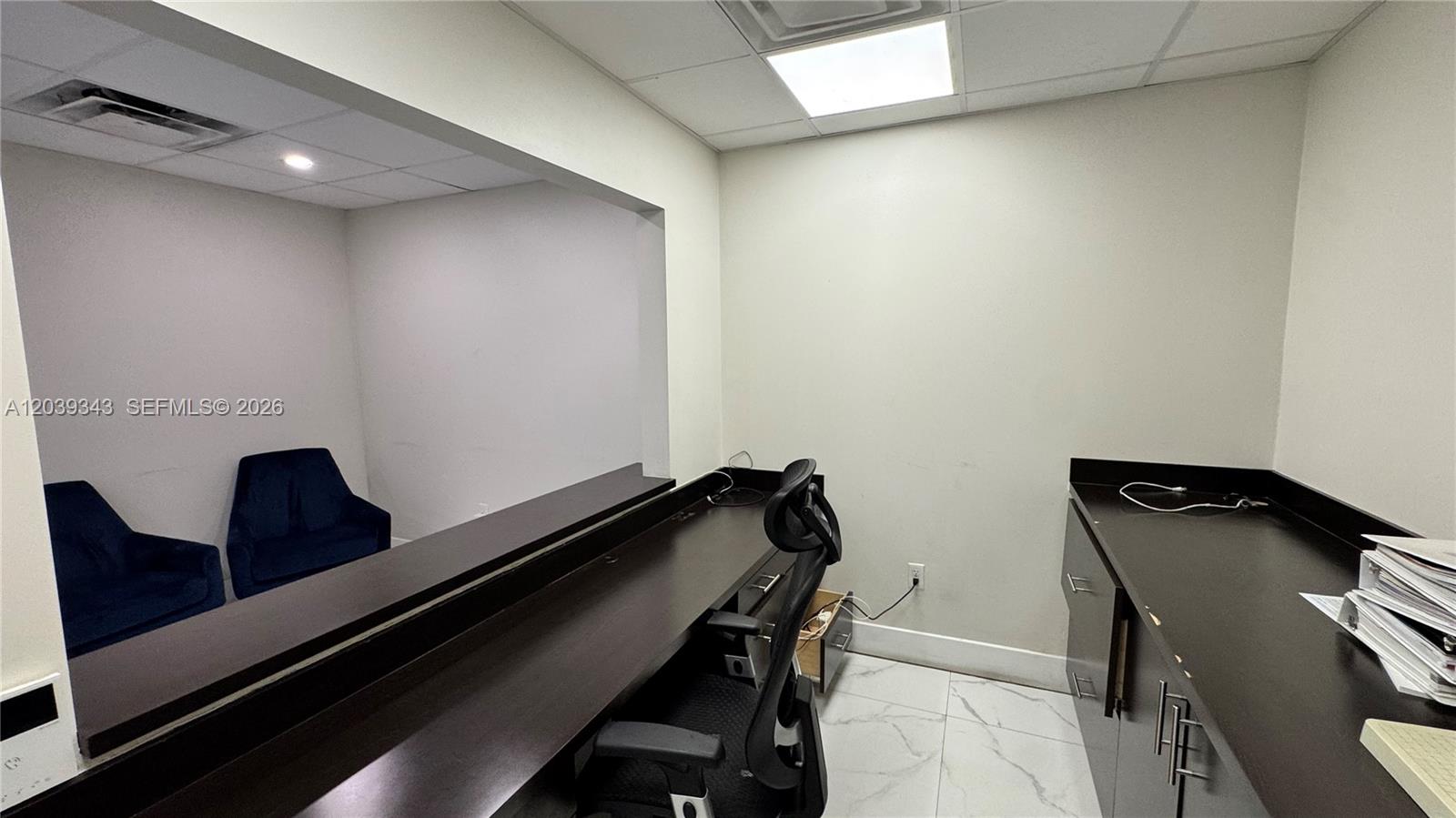 A small office space features a long dark desk, a rolling chair, and two blue chairs — MIAMI, FL