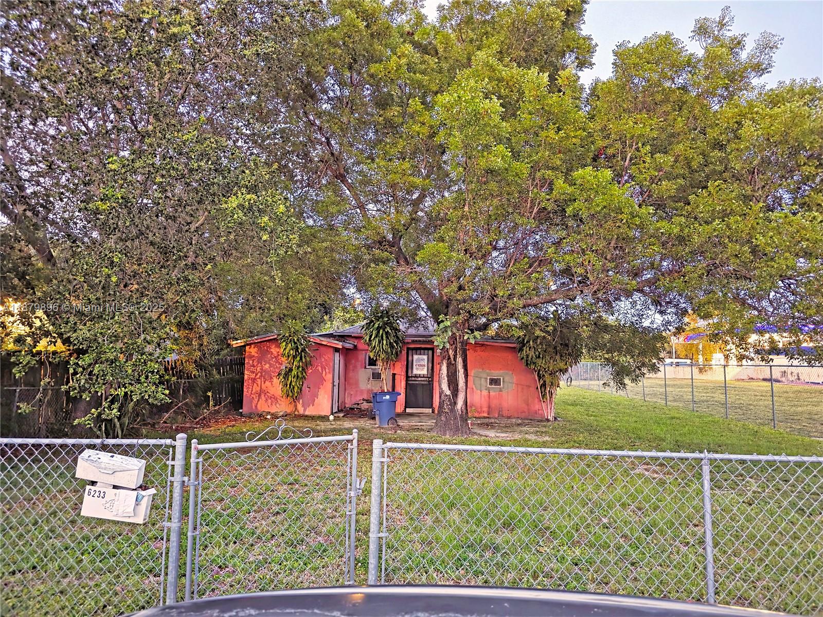 A small, red, single-story house sits behind a chain-link fence under large trees — 6233 NW 22nd Ct, Miami FL 33147, MIAMI, FL