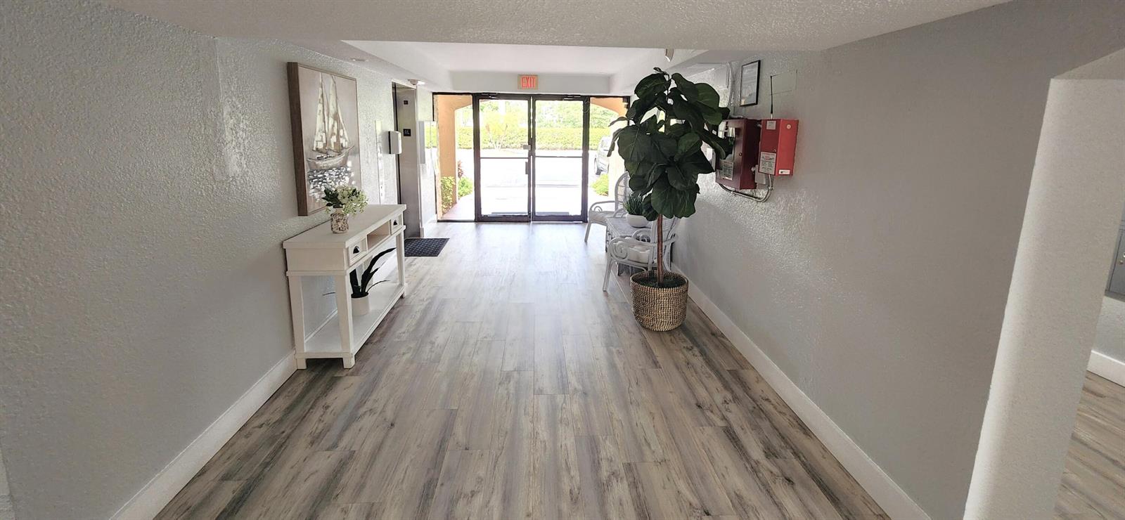 A bright building lobby features wood-look flooring, a white console table, and a large plant — 2455 Lindell Boulevard # 3511, Delray Beach FL 33444, DELRAY BEACH, FL