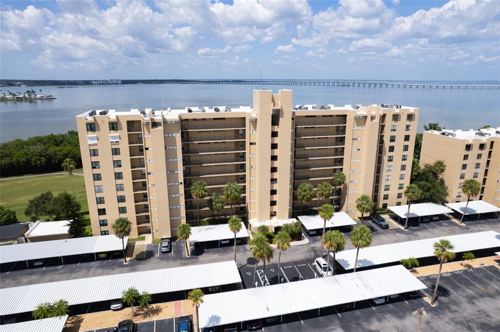 An aerial view shows a multi-story tan apartment building with covered parking and waterfront views — 2618 COVE CAY DRIVE UNIT 702, CLEARWATER, FL