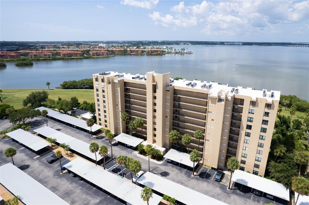 An aerial view shows a multi-story apartment building with covered parking lots near a lake — 2618 COVE CAY DRIVE UNIT 702, CLEARWATER, FL