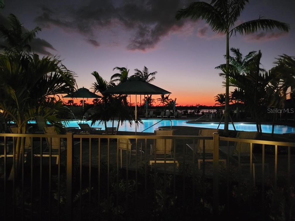 A swimming pool with lounge chairs and umbrellas is viewed through a fence at sunset — 17540 OPAL SAND DRIVE UNIT 204, VENICE, FL