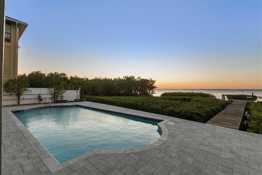 A rectangular swimming pool sits on a stone patio overlooking a bay at sunset — 3308 PALMA SOLA BOULEVARD, BRADENTON, FL