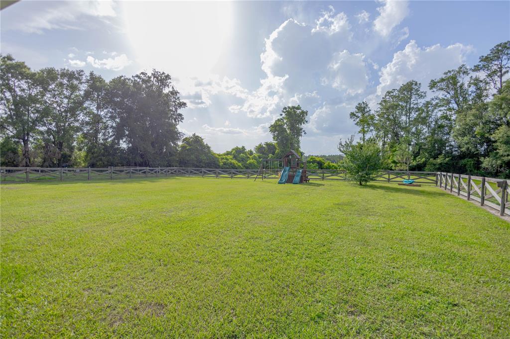 A large, fenced grassy backyard features a wooden playset with two slides under a sunny sky — 568 SW GERALD CONNER DRIVE, LAKE CITY, FL