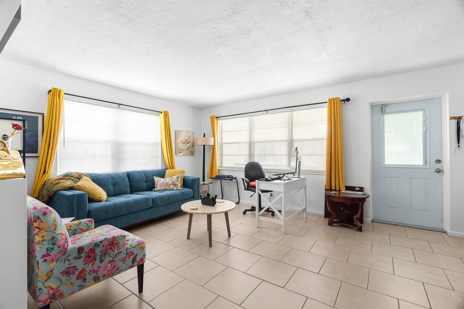 A living room with a blue sofa, floral armchair, desk, and tile flooring — 347 Windsor O, West Palm Beach FL 33417, WEST PALM BEACH, FL