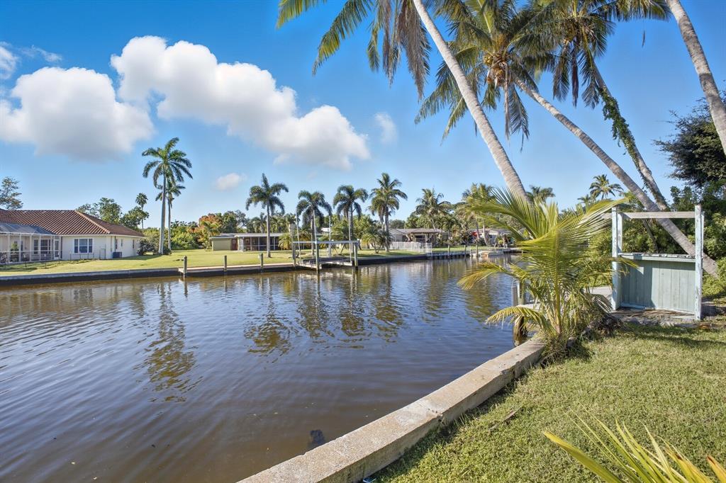 A canal view featuring a concrete seawall, palm trees, and residential homes in the background — 6915 9TH AVENUE NW, BRADENTON, FL