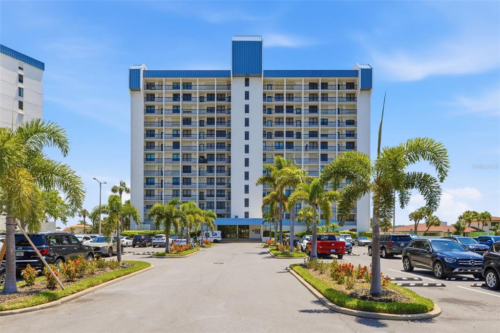 A multi-story residential building with balconies stands behind a parking lot with palm trees — 9415 BLIND PASS ROAD UNIT 806, ST PETE BEACH, FL