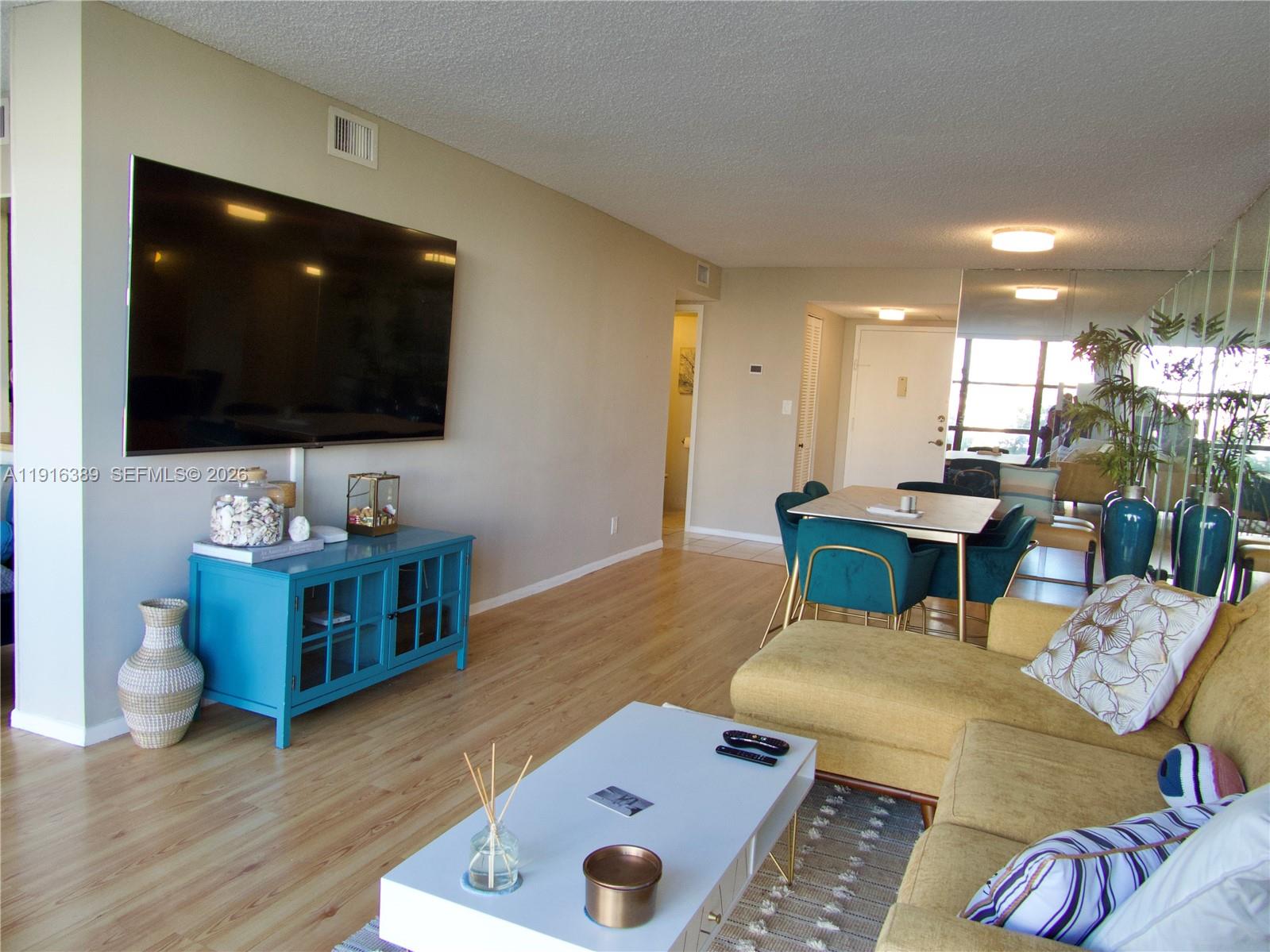 A living room features a wall-mounted television, a blue media console, and a yellow sectional — 800 Parkview Dr # 402, Hallandale Beach FL 33009, HALLANDALE BEACH, FL