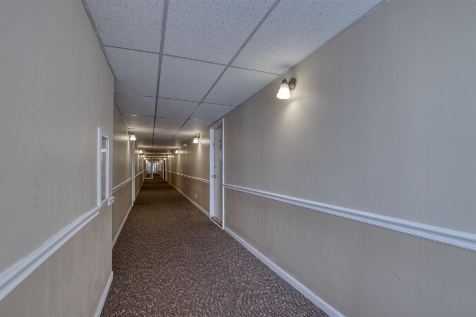 A long, narrow indoor hallway features beige walls, white trim, carpeted flooring, and ceiling tiles — 4657 Fountains Drive S # 206, FL 33467, FL