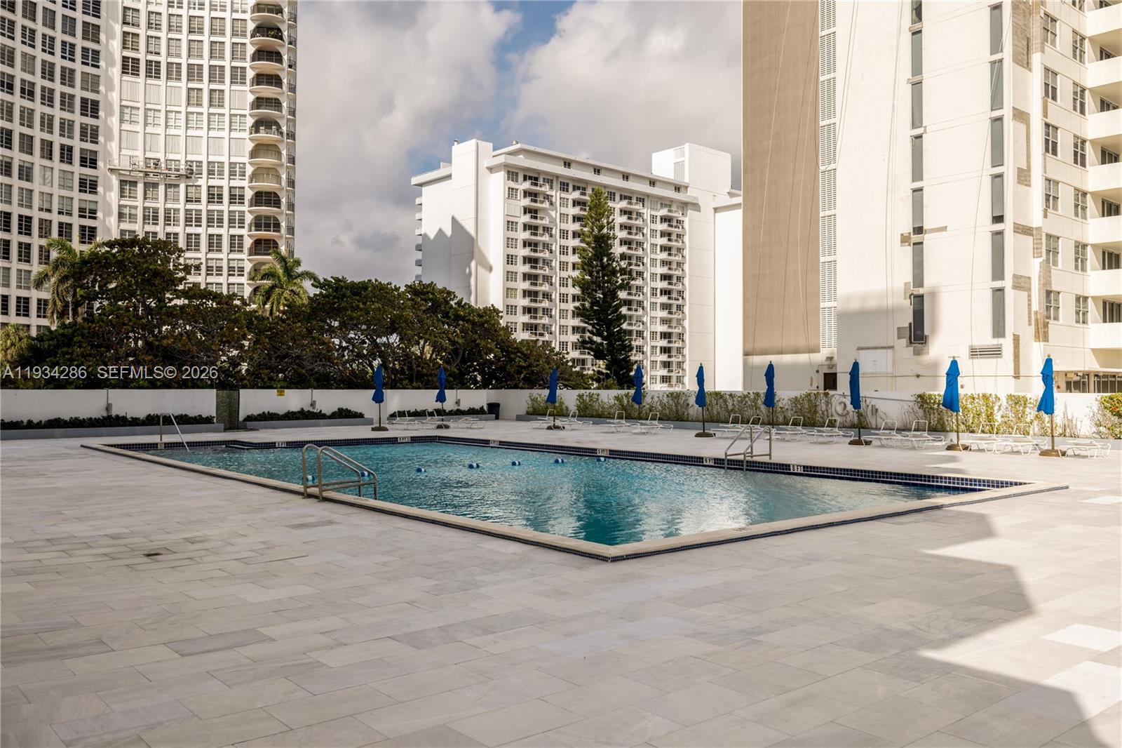An outdoor swimming pool surrounded by a tiled deck and multiple blue umbrellas — 5601 Collins Ave # 1019, Miami Beach FL 33140, MIAMI BEACH, FL