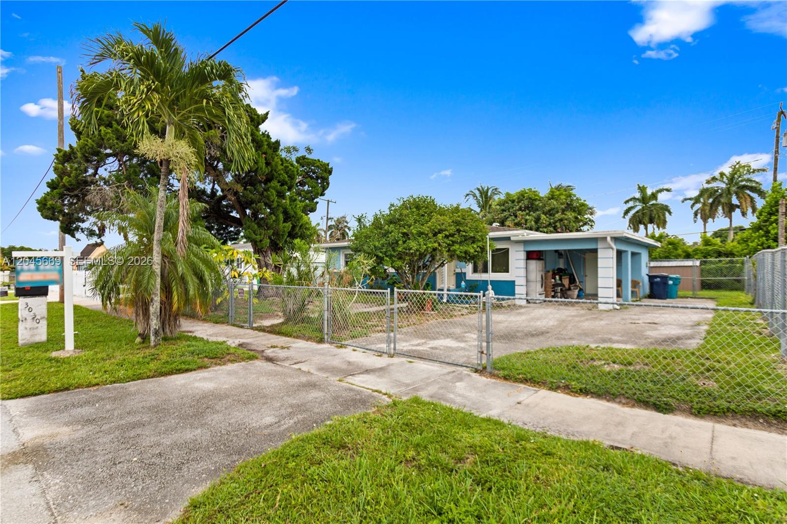 A single-story house with a carport sits behind a chain-link fence and palm trees — 3030 NW 176th St, Miami Gardens FL 33056, MIAMI GARDENS, FL