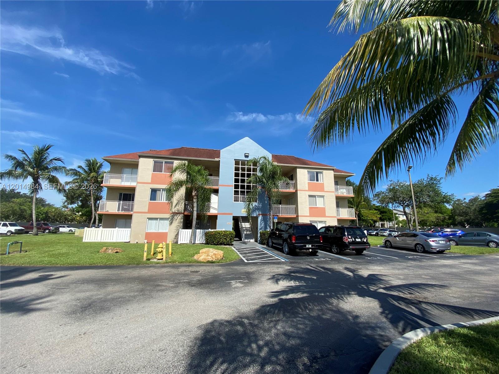 A three-story apartment building with a blue central facade stands behind a parking lot — 18820 NW 57th Ave # 203, Hialeah FL 33015, HIALEAH, FL