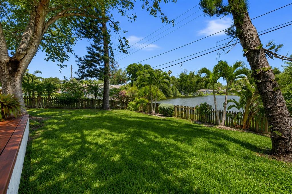 A grassy backyard features a wooden deck, palm trees, and a fence bordering a lake — 1918 BYRAM DRIVE, CLEARWATER, FL