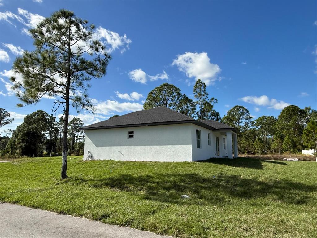 A side view of a white stucco house with a dark shingled roof and trees — 1121 FLORENCE STREET E, LEHIGH ACRES, FL