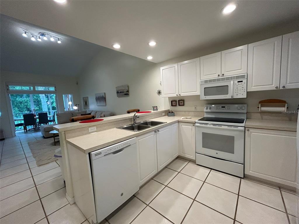 A kitchen with white cabinets, white appliances, and tile flooring overlooks an adjacent living area — 3605 54TH DRIVE W UNIT 203, BRADENTON, FL
