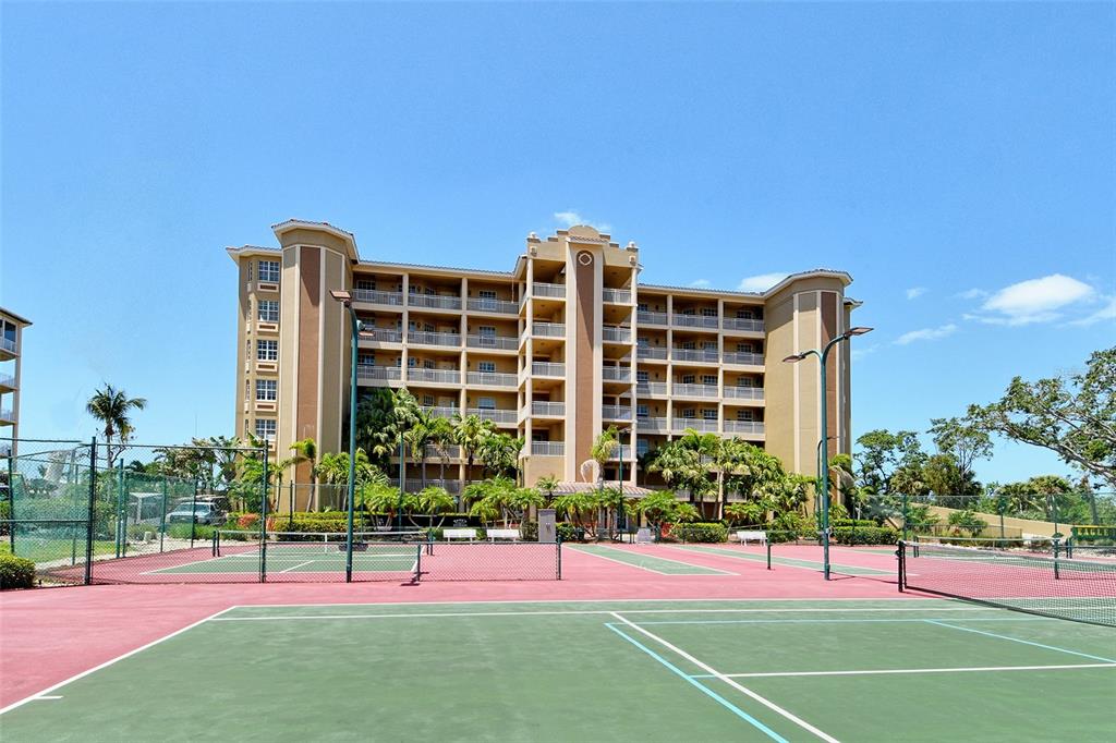A multi-story tan apartment building stands behind several outdoor tennis courts on a sunny day — 5100 JESSIE HARBOR DRIVE UNIT 504, OSPREY, FL