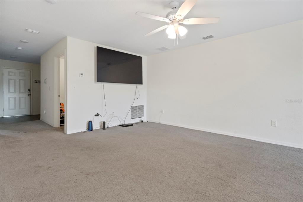 A living room features a wall-mounted television, gray carpeting, and a ceiling fan — 27 MAGNOLIA BLOSSOM LANE, KISSIMMEE, FL
