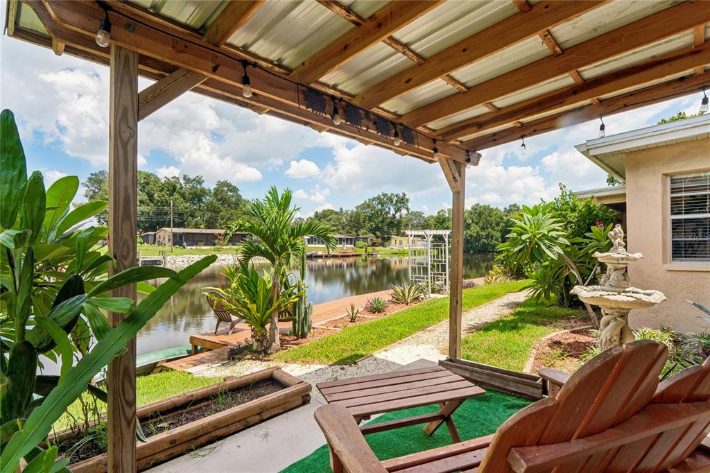 A covered patio overlooks a lake with a wooden deck, fountain, and tropical landscaping — 10681 SEMINOLE FOREST STREET E, SEMINOLE, FL